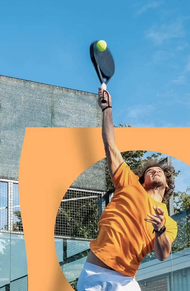 Man in oranje shirt slaat een bal met een padelracket tijdens een sprong op een buitenbaan.